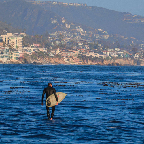 surfer with a surfboard walking into the ocean with a coastal town in the background