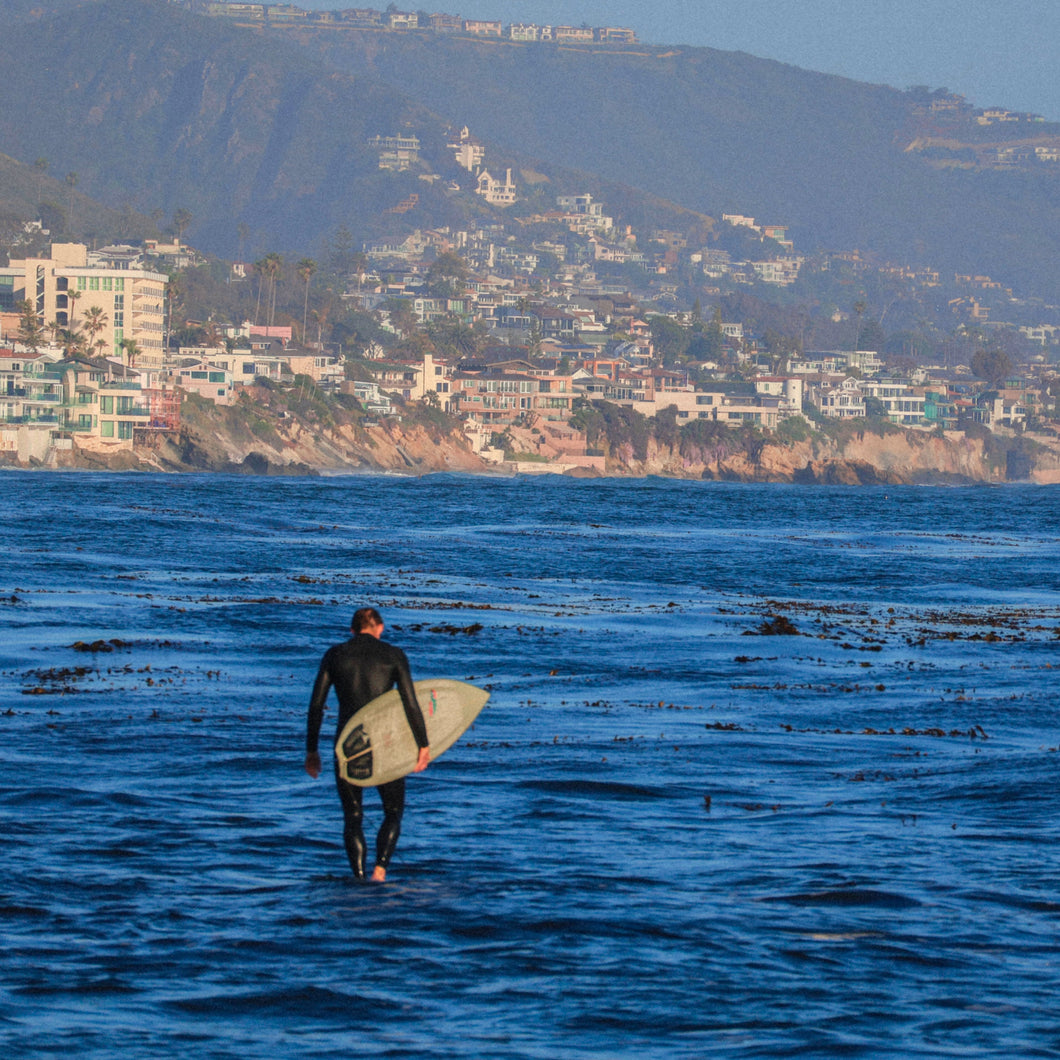 surfer with a surfboard walking into the ocean with a coastal town in the background