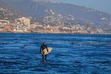 Person with a surfboard walking into the ocean with a coastal town in the background
