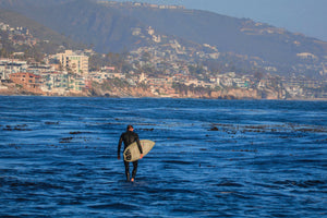 Person with a surfboard walking into the ocean with a coastal town in the background