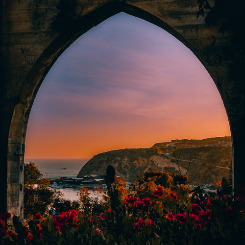 Sunset over a coastal landscape viewed through a stone archway with flowers in the foreground.