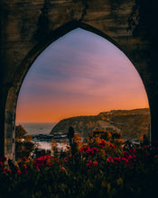 Sunset over a coastal landscape viewed through a stone archway with flowers in the foreground.