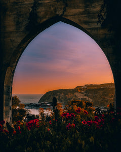 Sunset over a coastal landscape viewed through a stone archway with flowers in the foreground.
